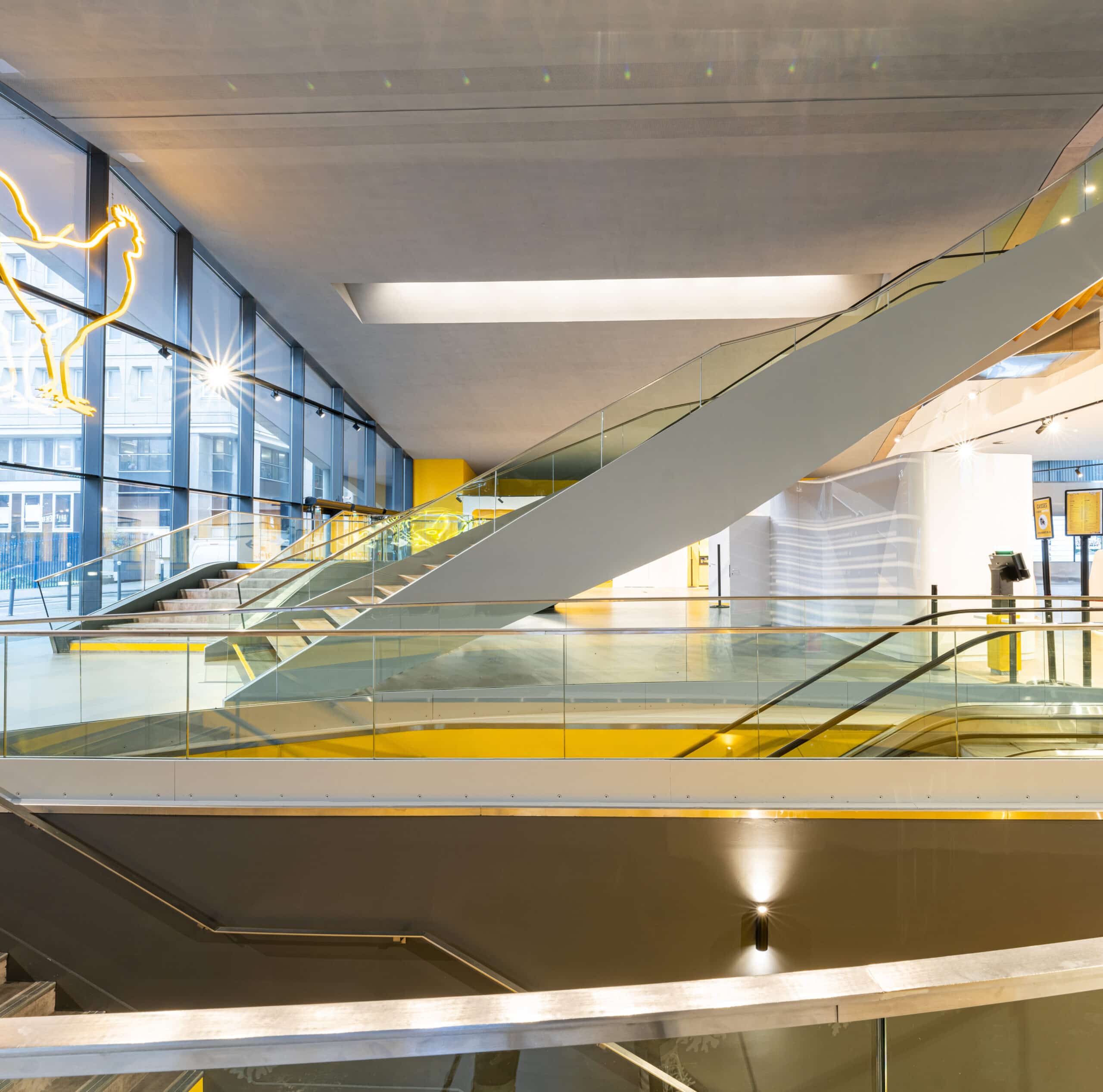 Espace lumineux du cinéma avec escalators modernes, garde-corps en verre et grande baie vitrée.