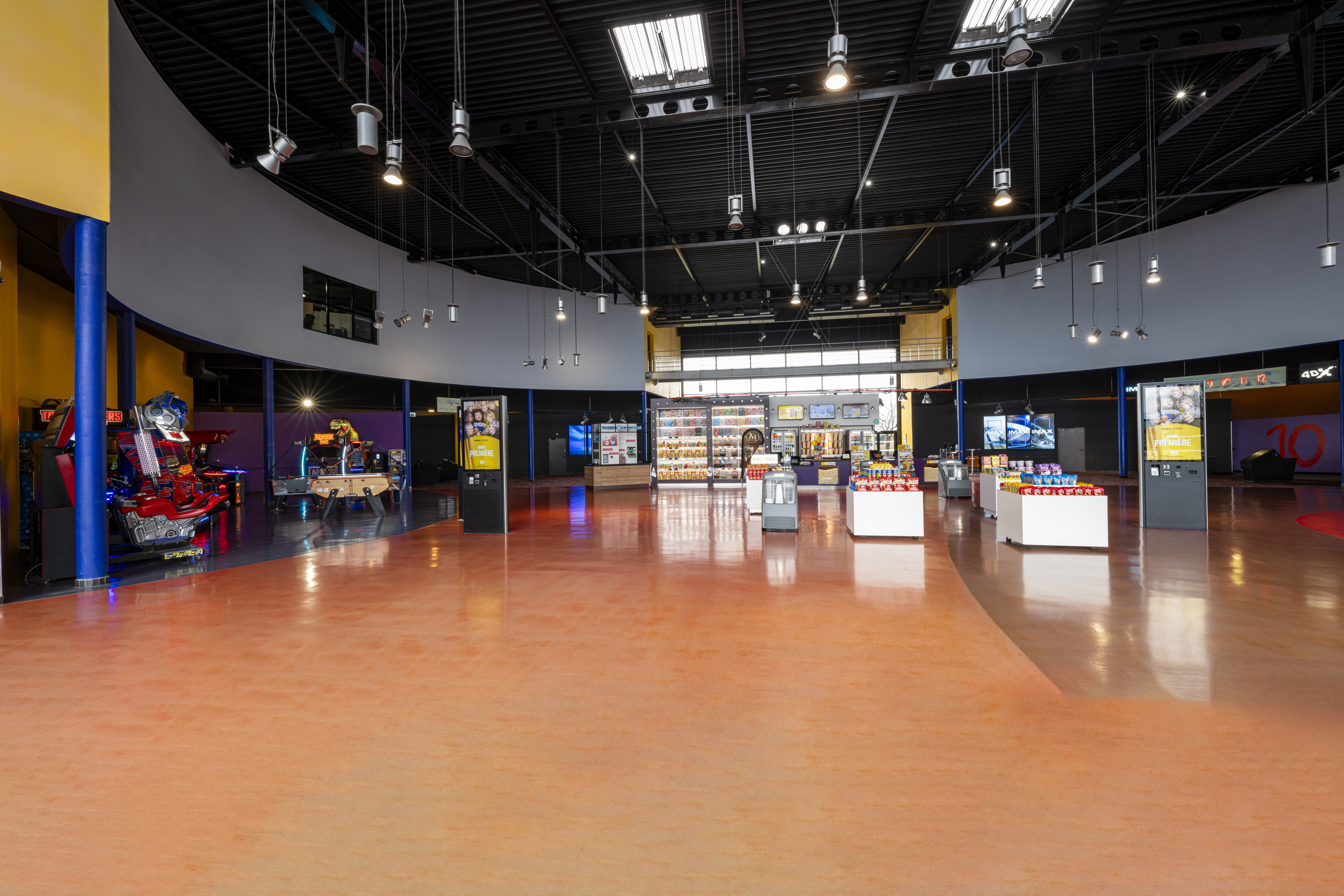 Grand hall moderne dans un cinéma avec un sol orange, des étagères de confiseries, des bornes interactives, et des jeux d'arcade en arrière-plan.