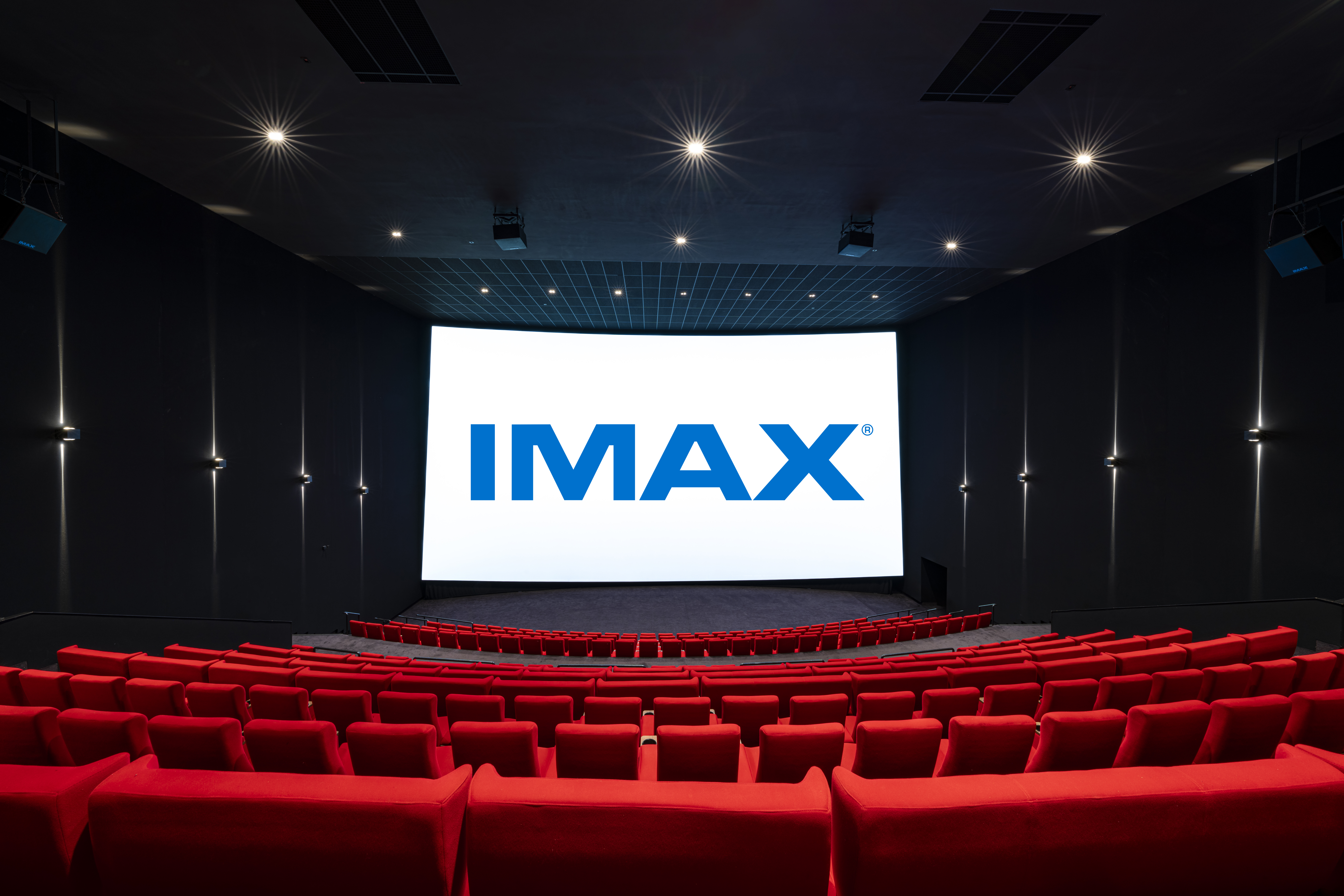 Vue de derrière d'une salle de cinéma équipée de la technologie IMAX montrant les fauteuils en velours rouges et l’agencement des rangées ainsi que son espace scénique.