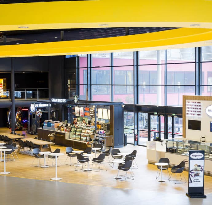 Espace lumineux avec un plafond jaune incurvé et une grande baie vitrée laissant passer la lumière naturelle. Au centre, un café Starbucks et un stand Häagen-Dazs sont entourés de tables blanches et de chaises noires modernes. L’arrière-plan montre des vitrines et des activités, créant une ambiance dynamique et conviviale.