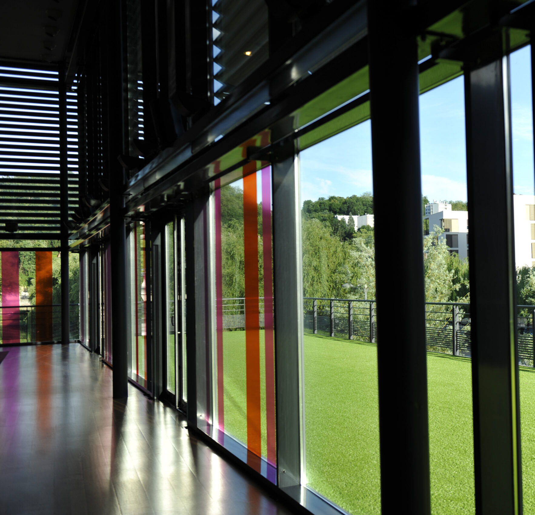 Vue intérieure sur une façade vitrée avec des bandes colorées verticales en orange, rose et violet. À travers les vitres, un espace extérieur est visible avec une pelouse verte et un paysage arboré. L'éclairage naturel illumine le sol sombre et l'ambiance est lumineuse et moderne.