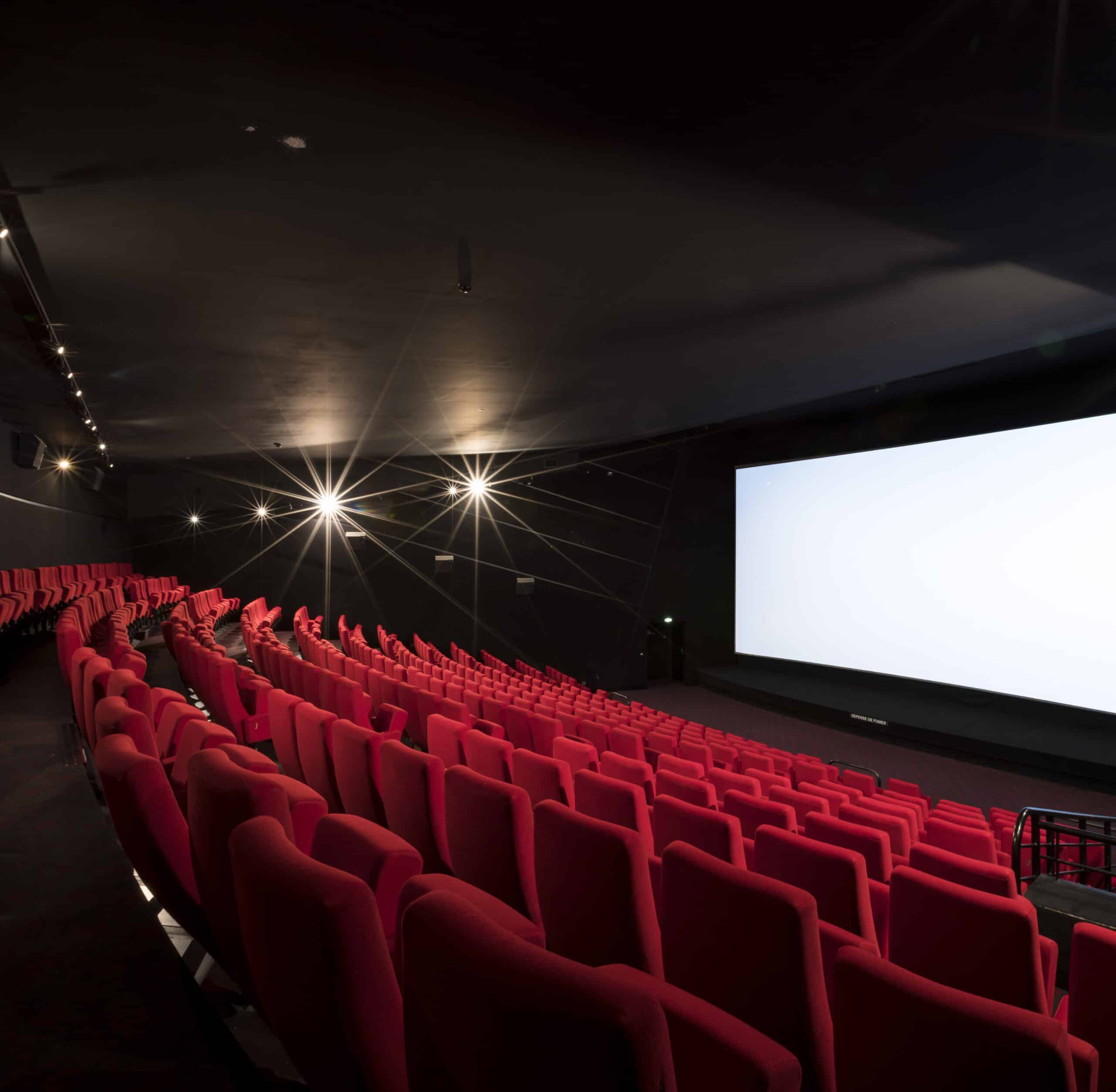 Vue depuis l’arrière d'une salle de cinéma avec ses rangées de fauteuils en velours rouges orientés vers l’écran.
