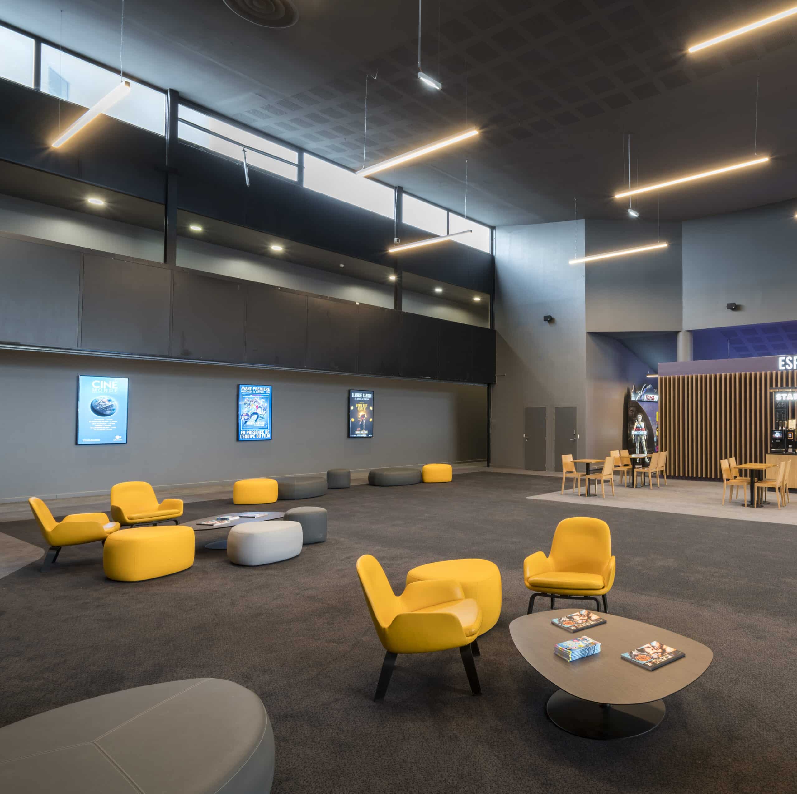 Espace lounge du cinéma avec fauteuils jaunes, poufs gris et tables modernes sur moquette sombre. Zone d’affiches lumineuses le long du mur et coin jeux visible à droite. Éclairage linéaire suspendu et plafond haut accentuent le design spacieux.