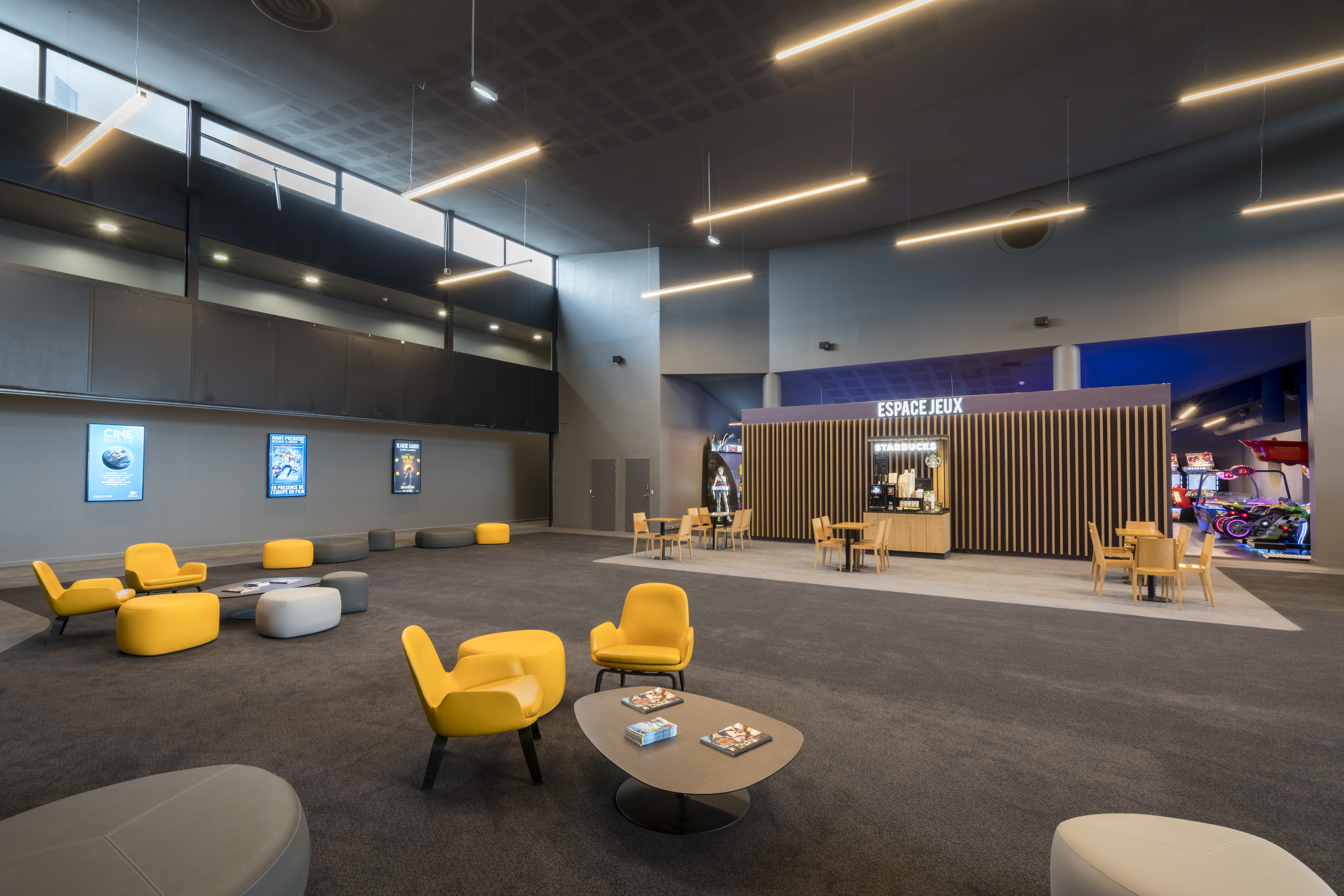 Espace lounge du cinéma avec fauteuils et poufs jaunes et gris sur moquette sombre, tables en bois et coin café Starbucks intégré. Bornes d’arcade lumineuses visibles à droite, murs décorés d’affiches de cinéma, et éclairage moderne au plafond.