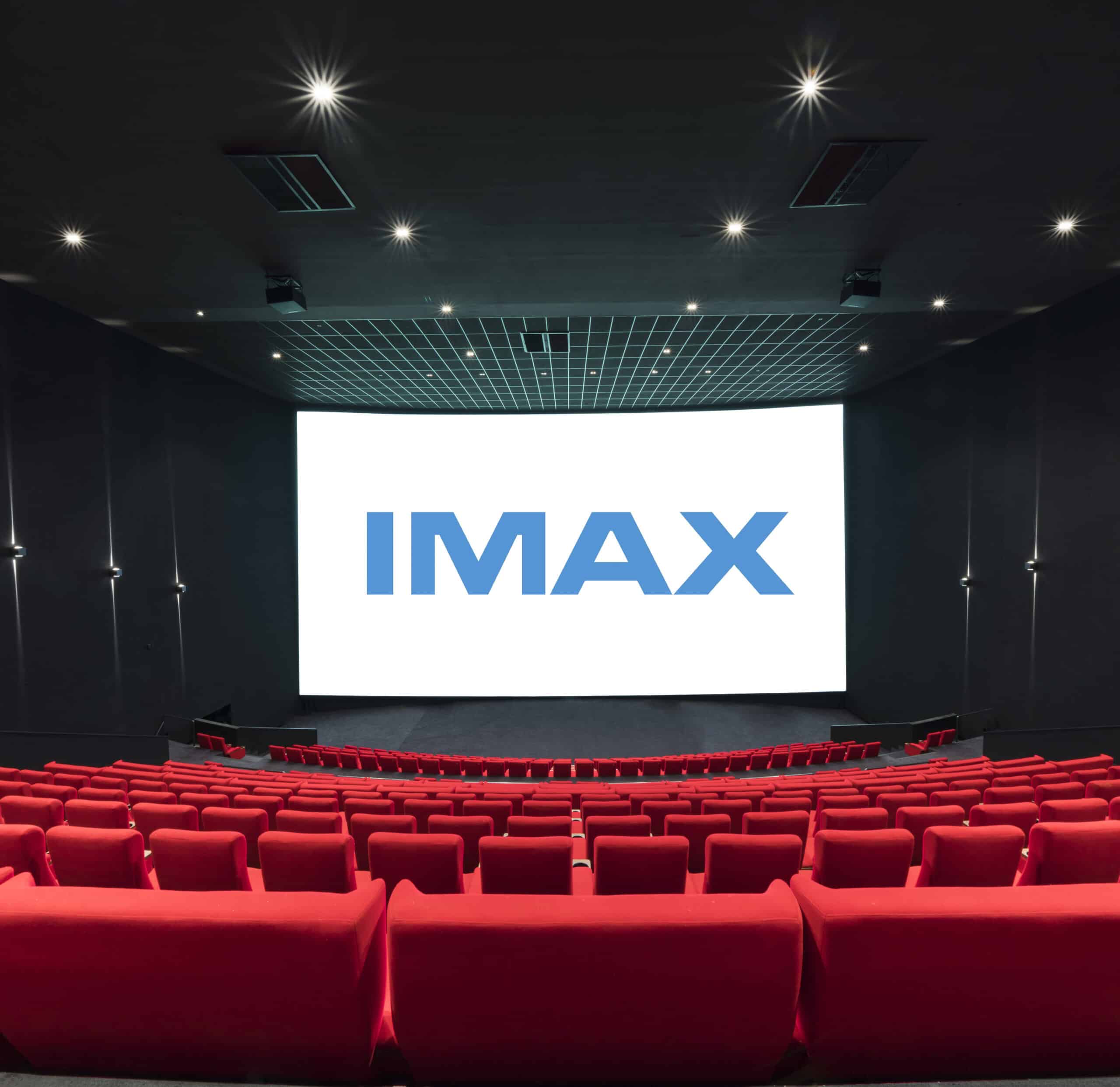 Vue de derrière d'une salle de cinéma équipée de la technologie IMAX montrant les fauteuils en velours rouges et l’agencement des rangées ainsi que son espace scénique.