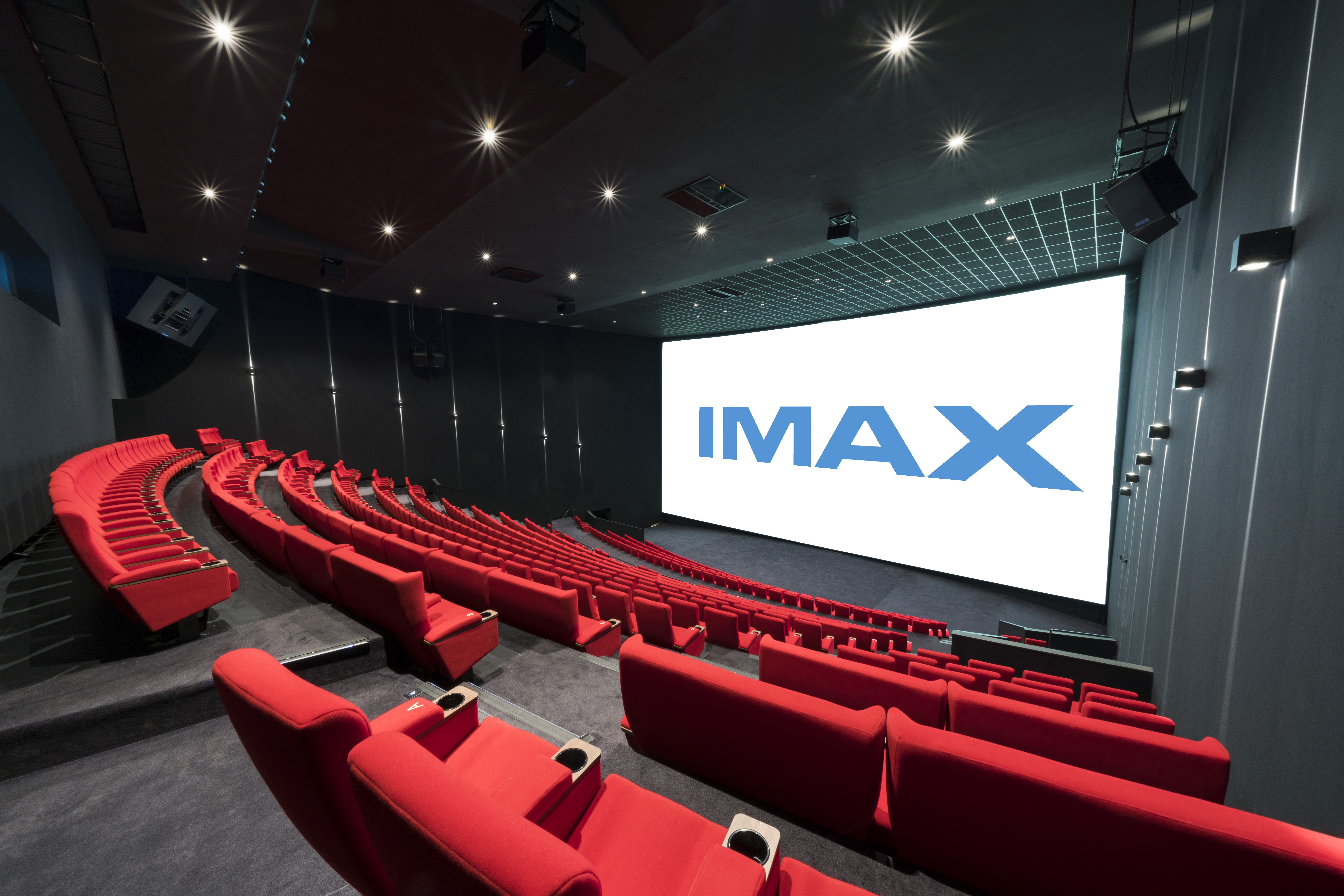 Vue de derrière d'une salle de cinéma équipée de la technologie IMAX montrant les fauteuils en velours rouges et l’agencement des rangées ainsi que son espace scénique.