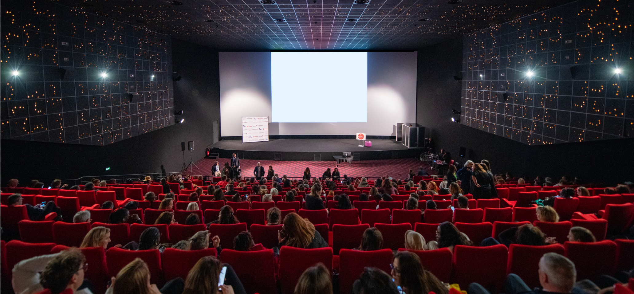 Salle de cinéma remplie de spectateurs, vue de derrière, lors d’un événement professionnel de type conférence.