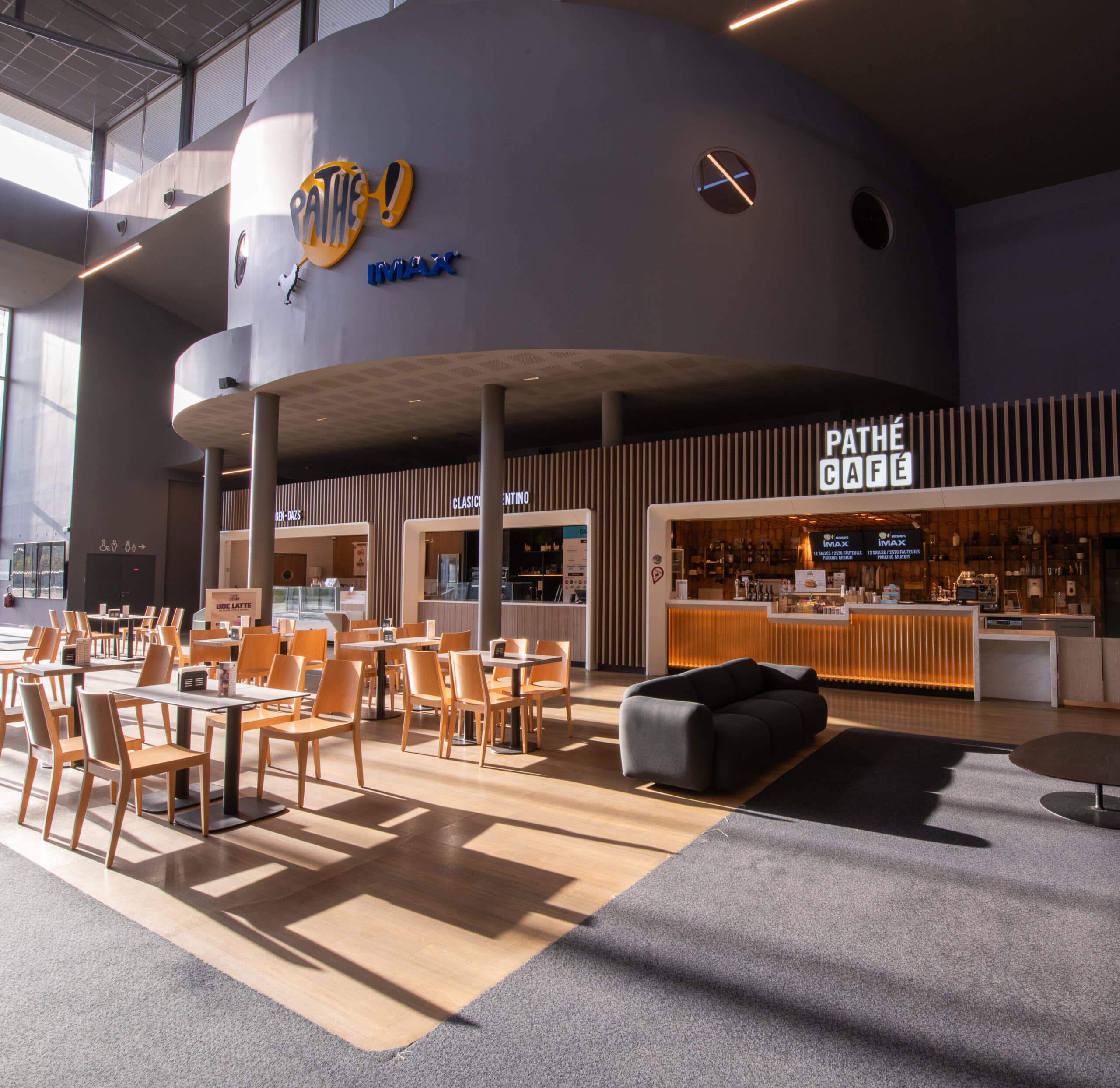 Hall du cinéma Pathé avec enseigne lumineuse Pathé IMAX sur un mur incurvé gris, comptoirs Häagen-Dazs, Classico Argentino et Pathé Café. Espace au design moderne avec tables et chaises en bois, canapé noir et sols combinant moquette et parquet. Lumière naturelle pénétrant par les grandes baies vitrées.