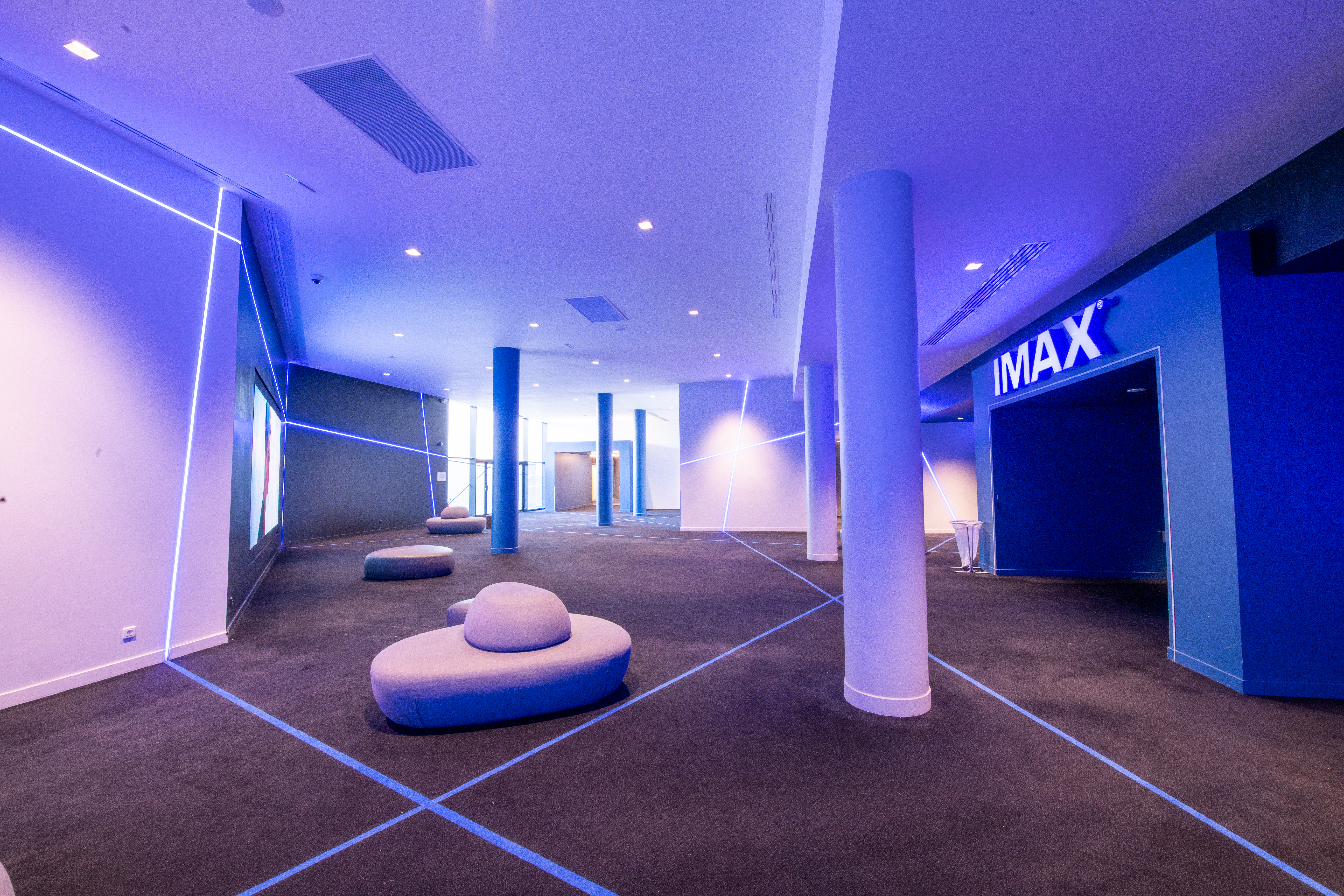 Couloir menant à l’entrée IMAX avec ambiance lumineuse bleue, sol en moquette sombre et sièges arrondis modernes. Décoration composée de lignes lumineuses sur les murs et colonnes blanches. Baies vitrées au fond offrant une lumière naturelle.