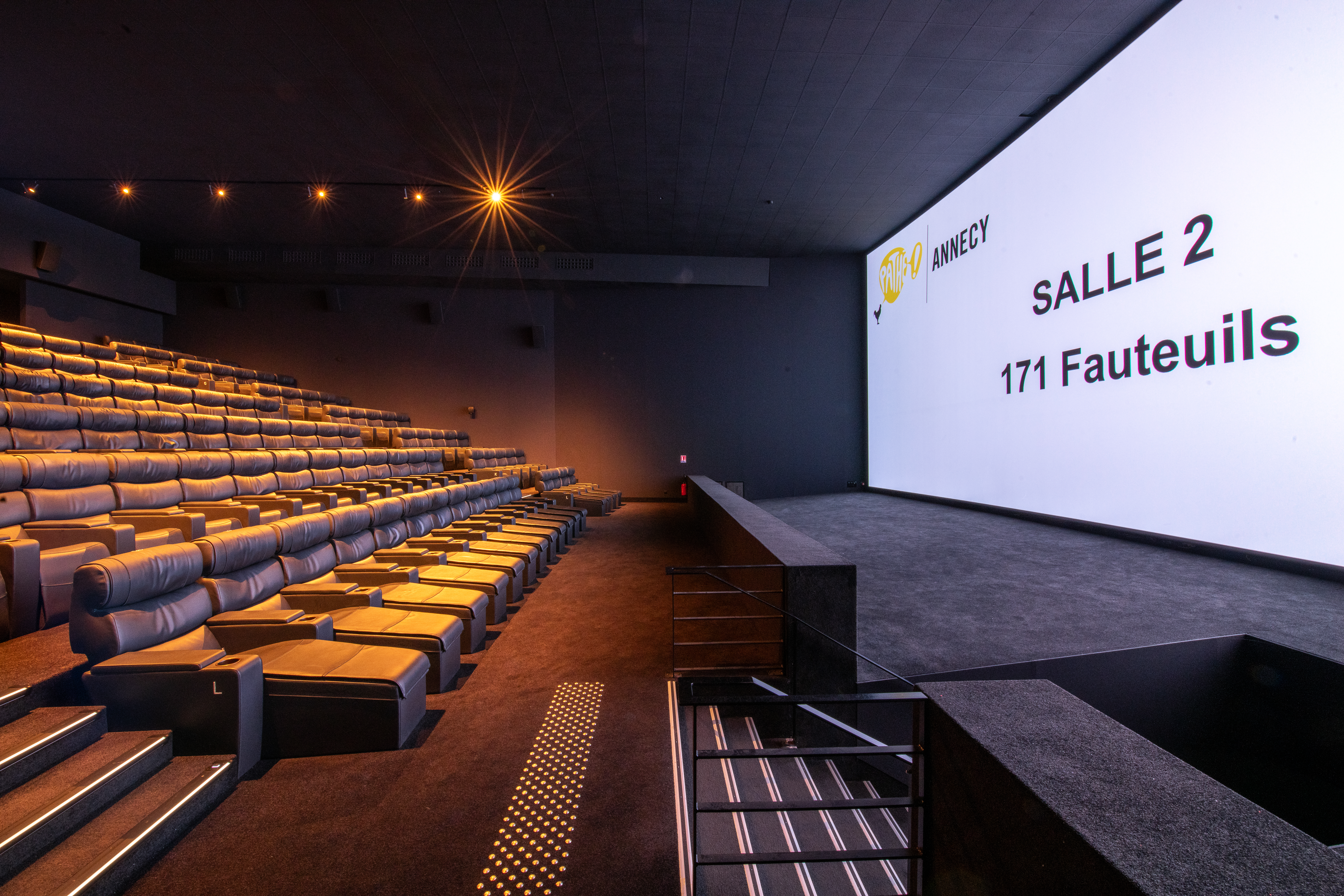 Vue latérale de la salle 2 du cinéma Pathé Annecy montrant les fauteuils en velours rouges et l’agencement des rangées ainsi qu'une partie de son espace scénique.