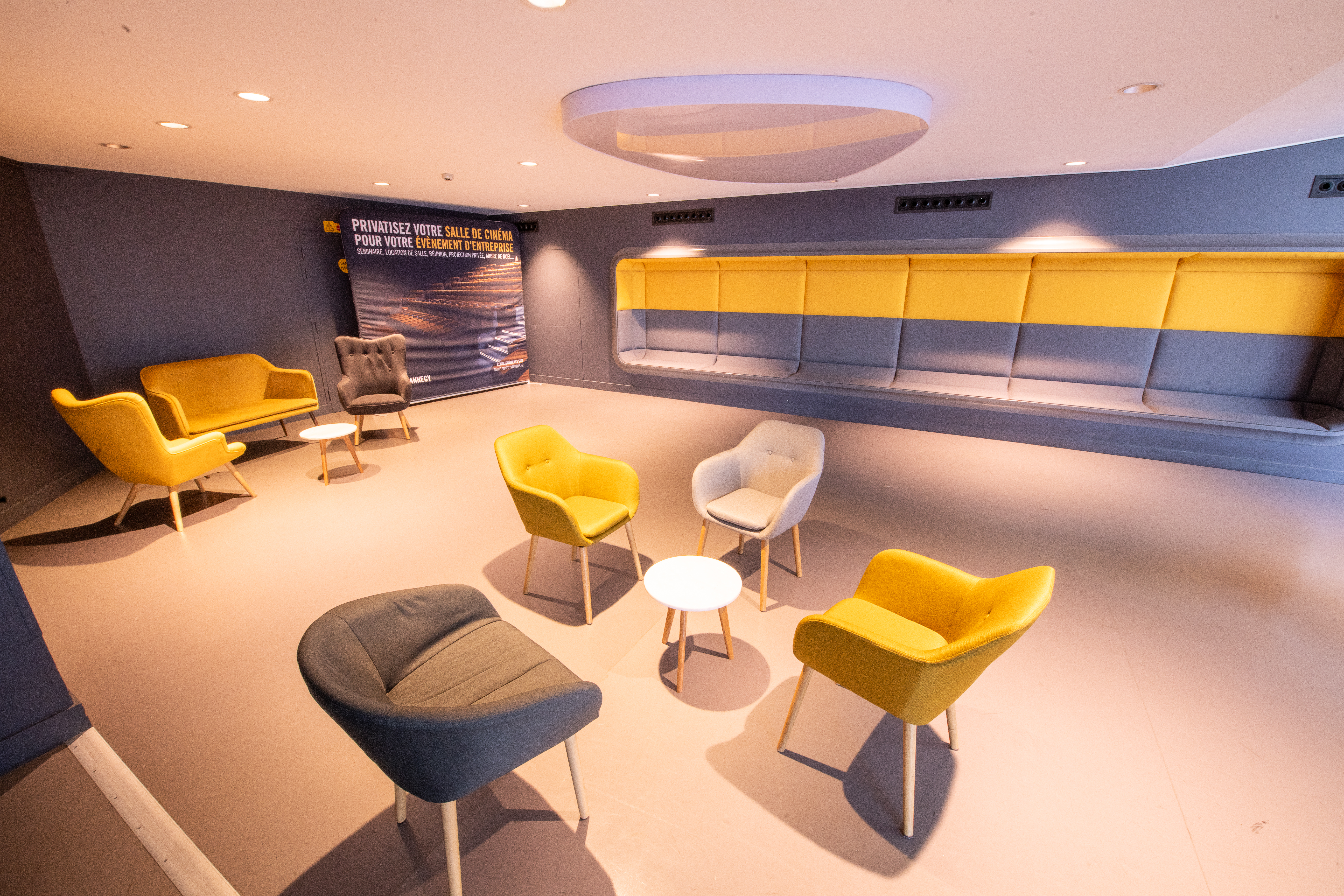 Salon d’attente au cinéma avec fauteuils et canapés jaune et gris, petites tables rondes blanches, et banquette murale intégrée. Décor lumineux avec mur gris foncé et affiche promotionnelle sur la location de salles pour événements.