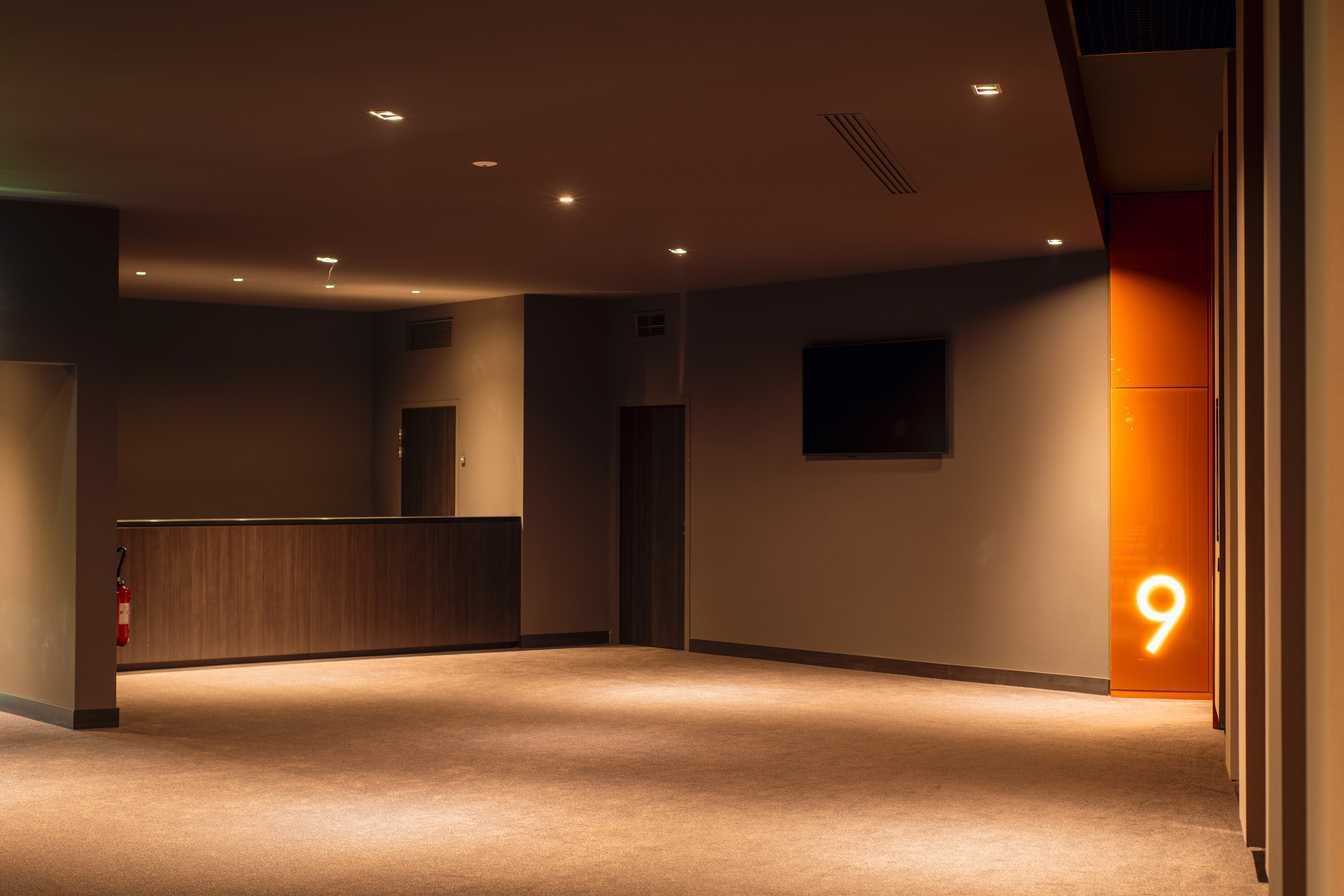 Espace d'attente sobre et chaleureux dans un cinéma, avec un éclairage doux, un écran mural et une porte numérotée 