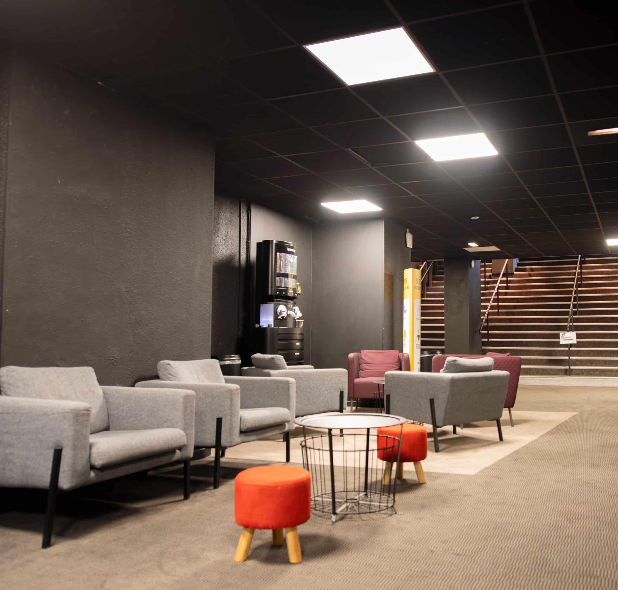 Espace de réception du cinéma avec fauteuils gris et rouges, tables basses et machine à café, près d’un escalier.