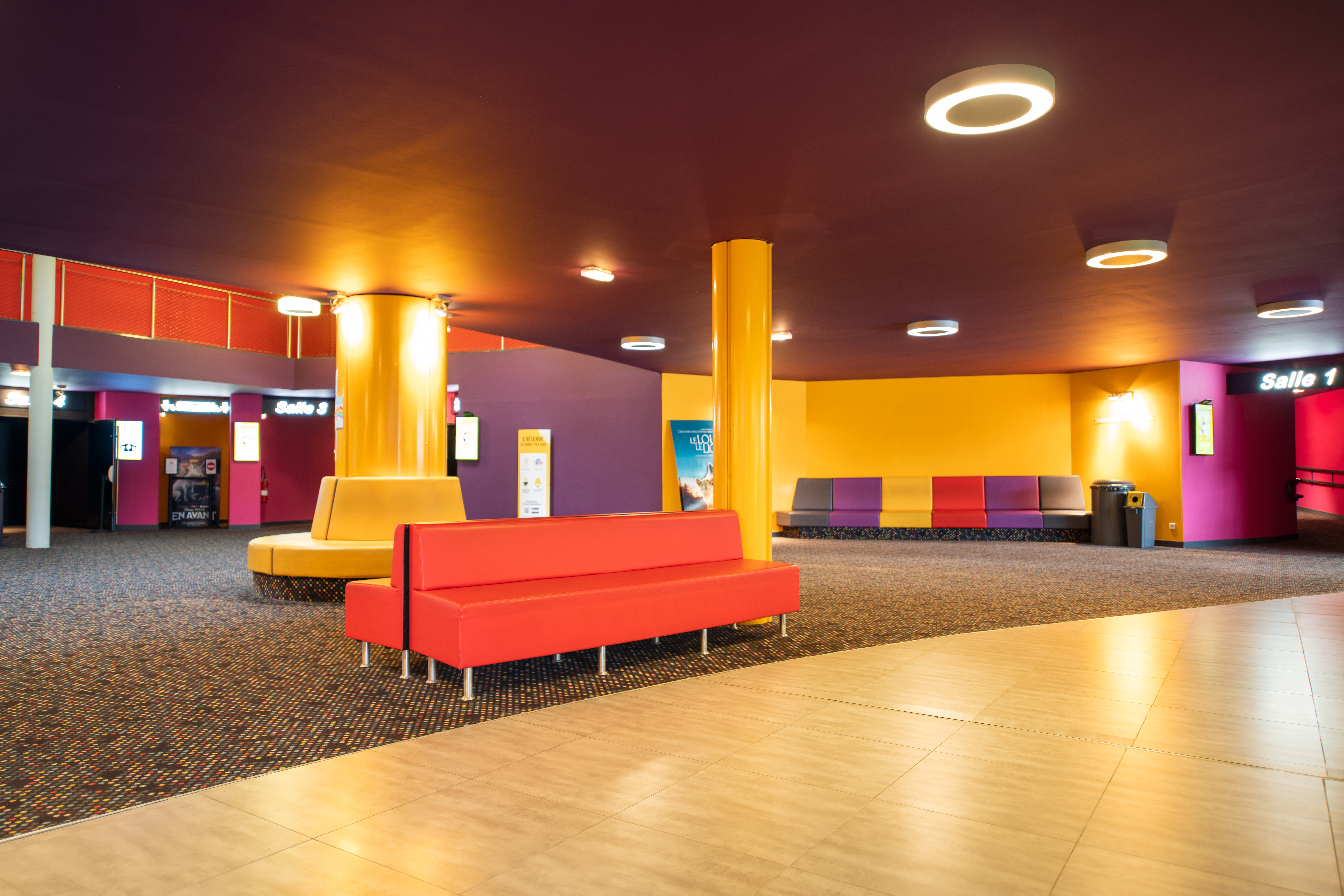 Espace de réception d'un cinéma avec des sièges colorés, un sol à motifs et des colonnes jaunes, entouré par des entrées de salles numérotées.