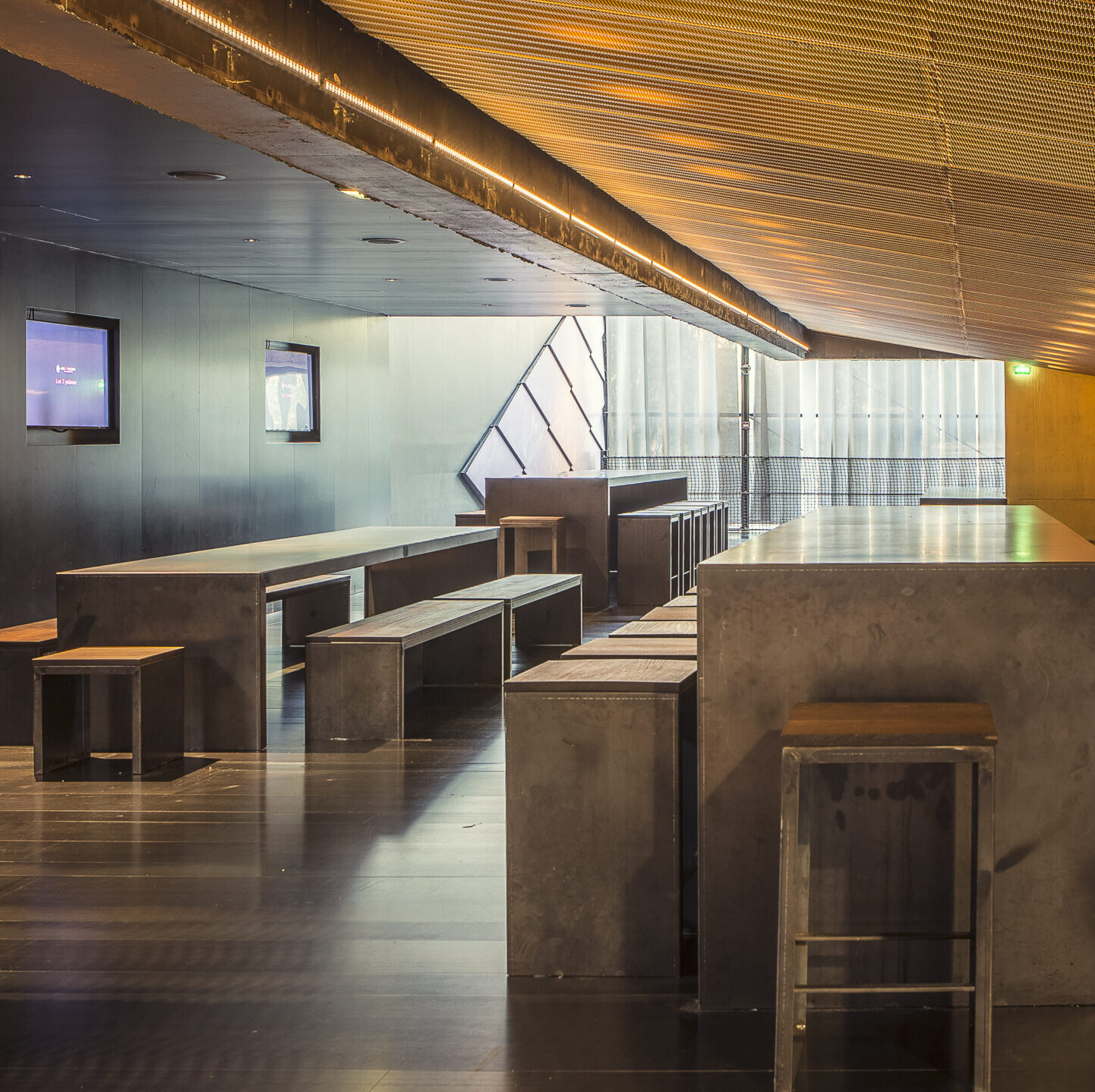Espace moderne et épuré avec tables et bancs en métal. Éclairage chaleureux au plafond, murs sombres et écrans sur les côtés. Grandes vitres apportant une lumière naturelle.