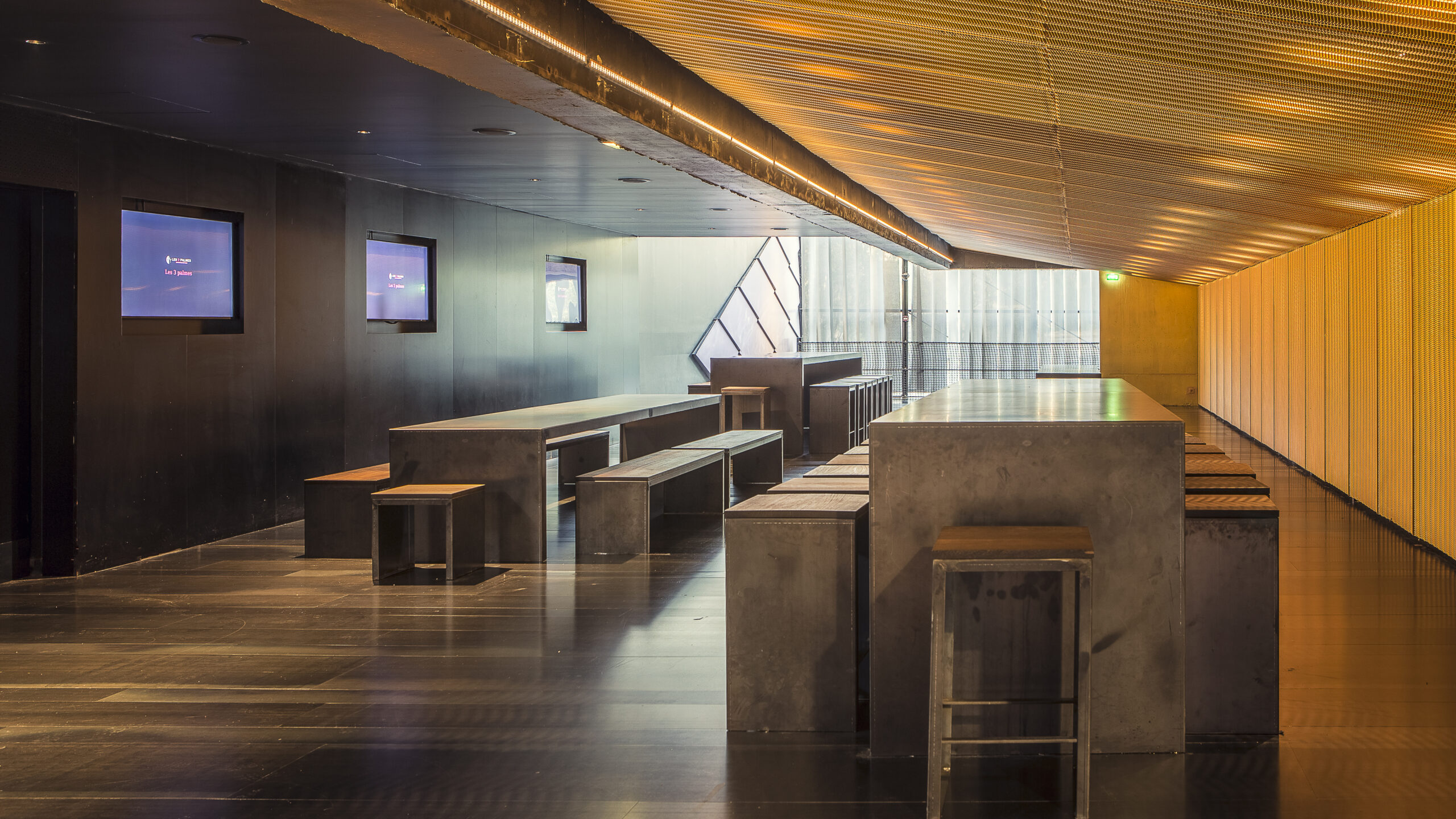 Espace moderne et épuré avec tables et bancs en métal. Éclairage chaleureux au plafond, murs sombres et écrans sur les côtés. Grandes vitres apportant une lumière naturelle.