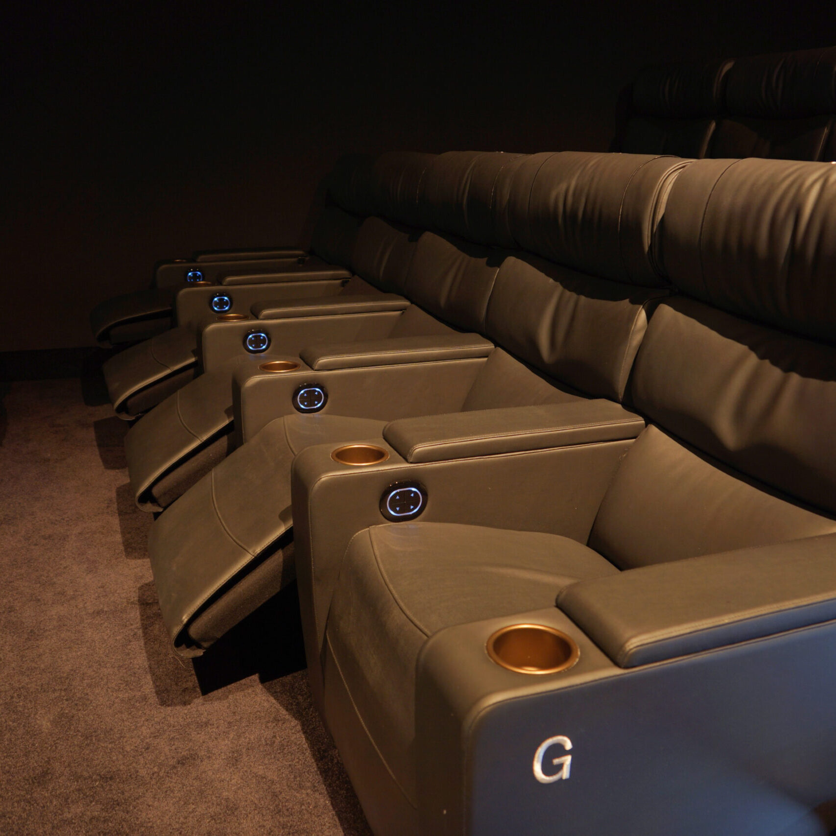 Rangée de fauteuils de cinéma en cuir marron foncé avec assise inclinable. Chaque fauteuil est équipé d’accoudoirs disposant d’un porte-gobelet doré et d’un bouton lumineux de commande. Le sol est recouvert de moquette sombre, et l’ambiance est tamisée.