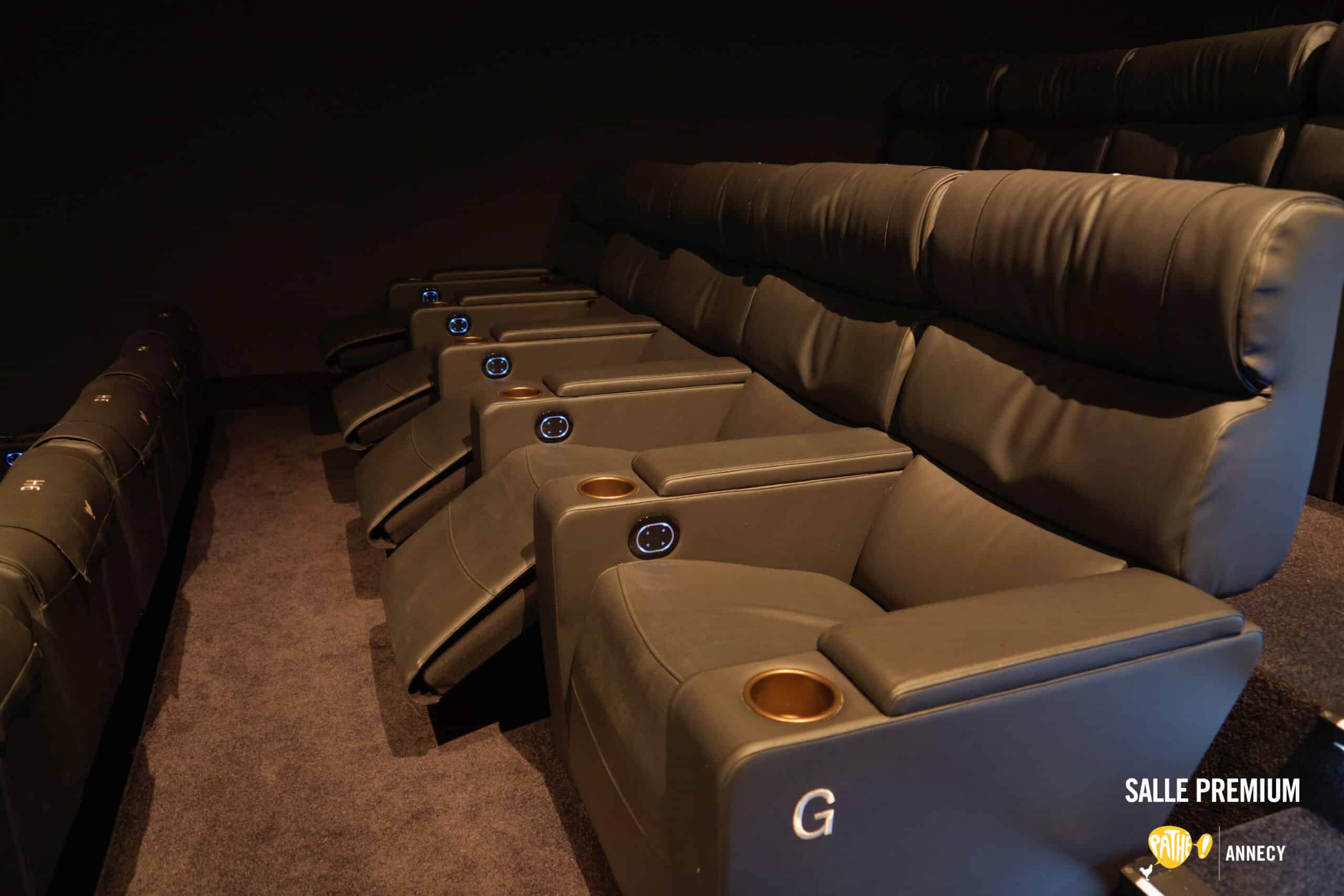Rangée de fauteuils de cinéma en cuir marron foncé avec assise inclinable. Chaque fauteuil est équipé d’accoudoirs disposant d’un porte-gobelet doré et d’un bouton lumineux de commande. Le sol est recouvert de moquette sombre, et l’ambiance est tamisée.