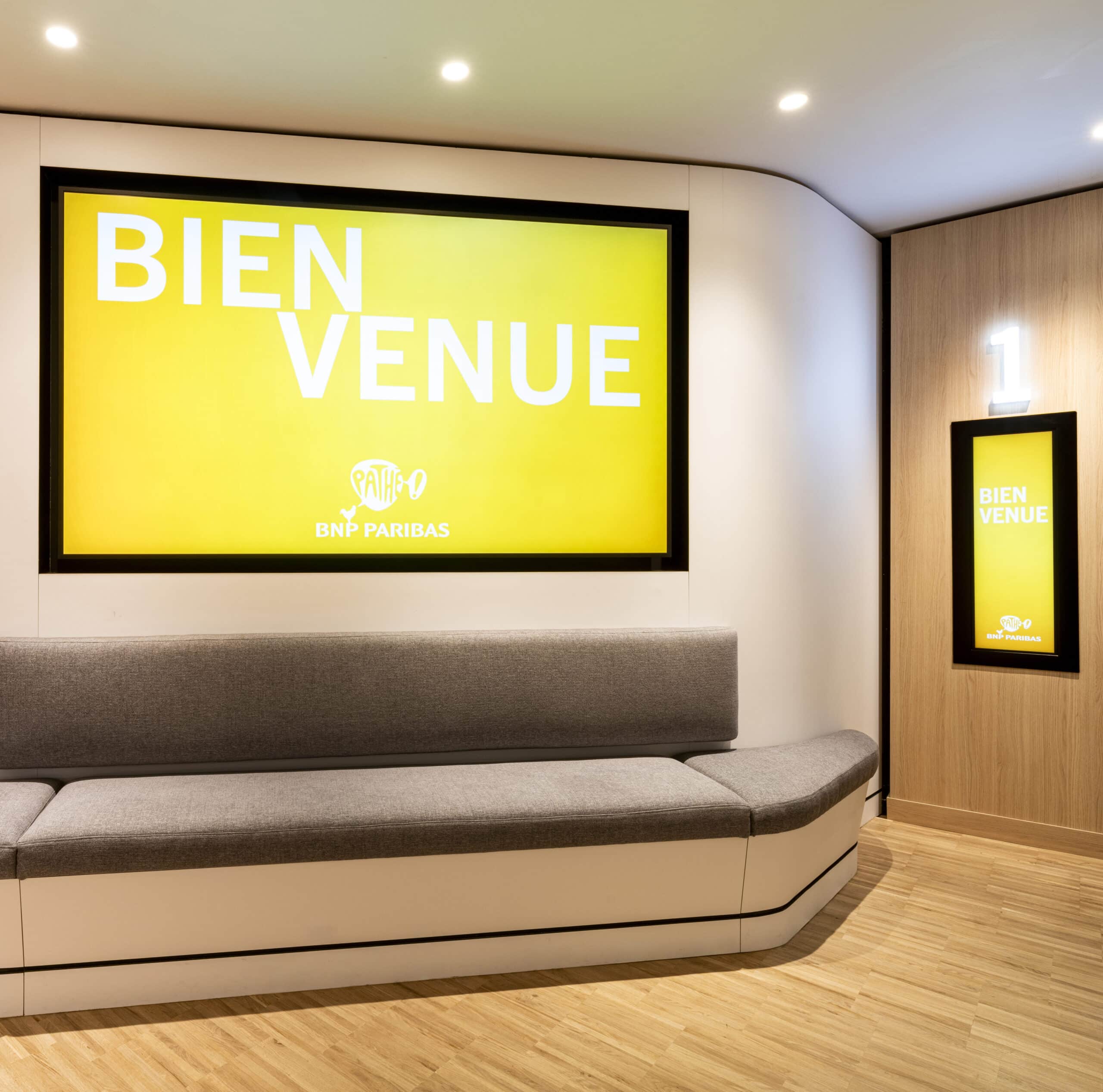 Espace accueil du cinéma avec écran lumineux « Bienvenue », banquette grise et signalétique moderne.
