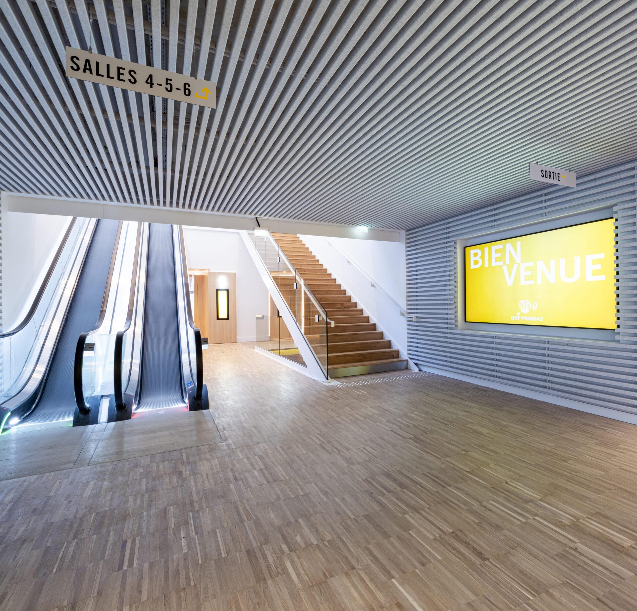 Espace circulatoire du cinéma avec escalators, escalier en bois et écran lumineux « Bienvenue »
