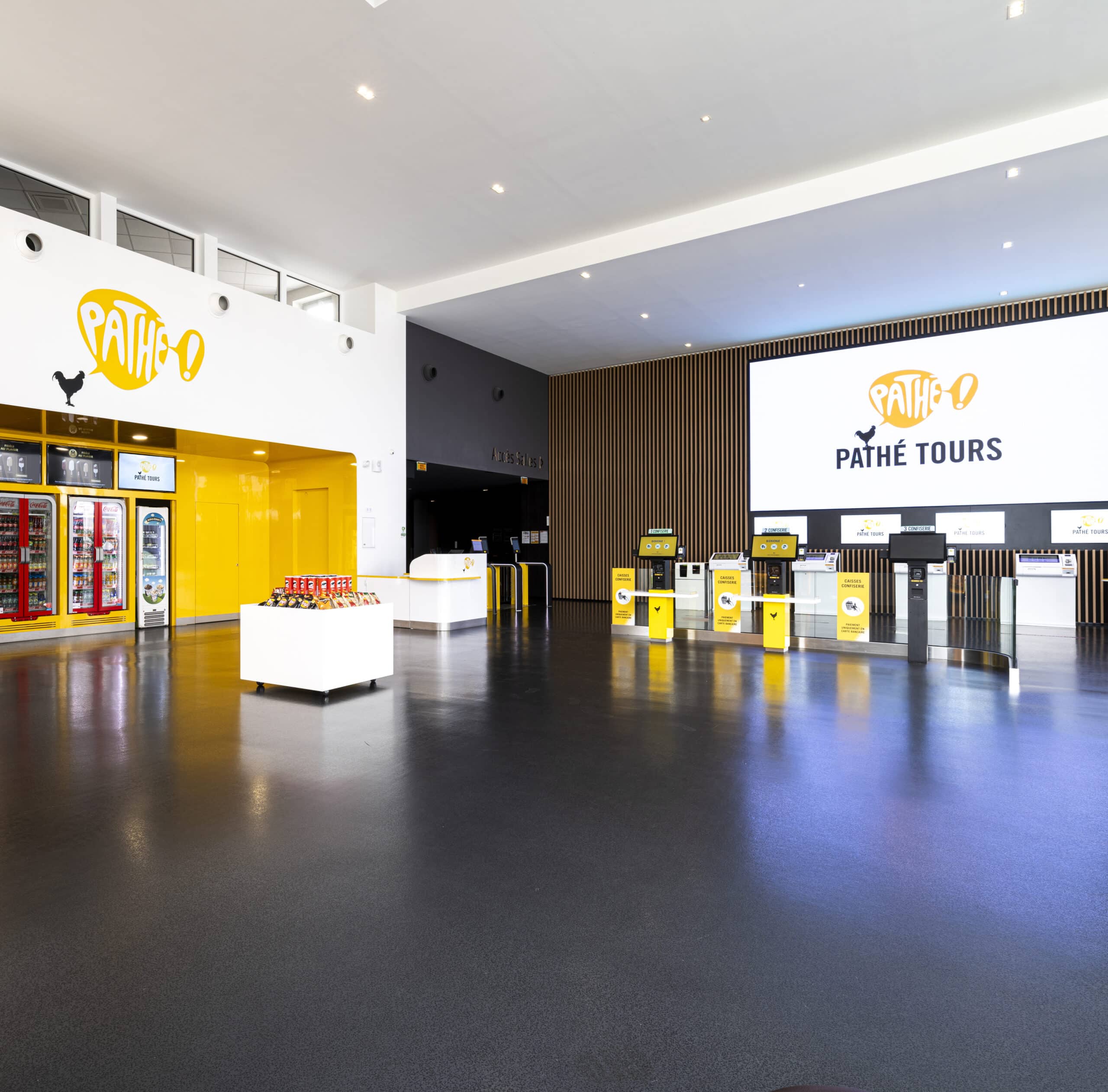 Hall d'entrée moderne du cinéma Pathé Tours avec des murs jaunes, des écrans lumineux, un snack-bar et des bornes automatiques pour l'achat de billets.