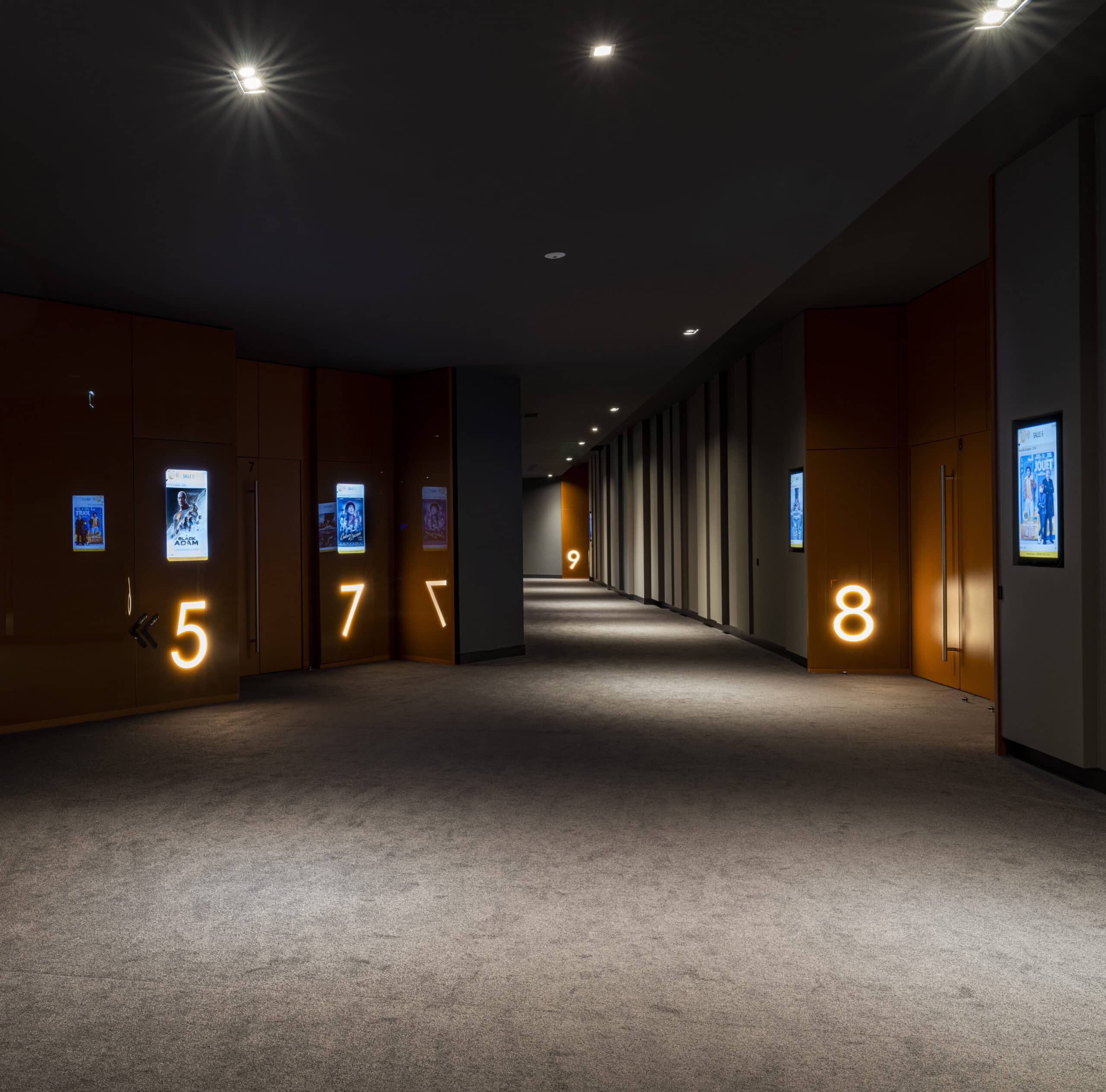 Couloir sombre et élégant dans un cinéma, avec des portes numérotées illuminées de 5 à 9 et des affiches de films éclairées.