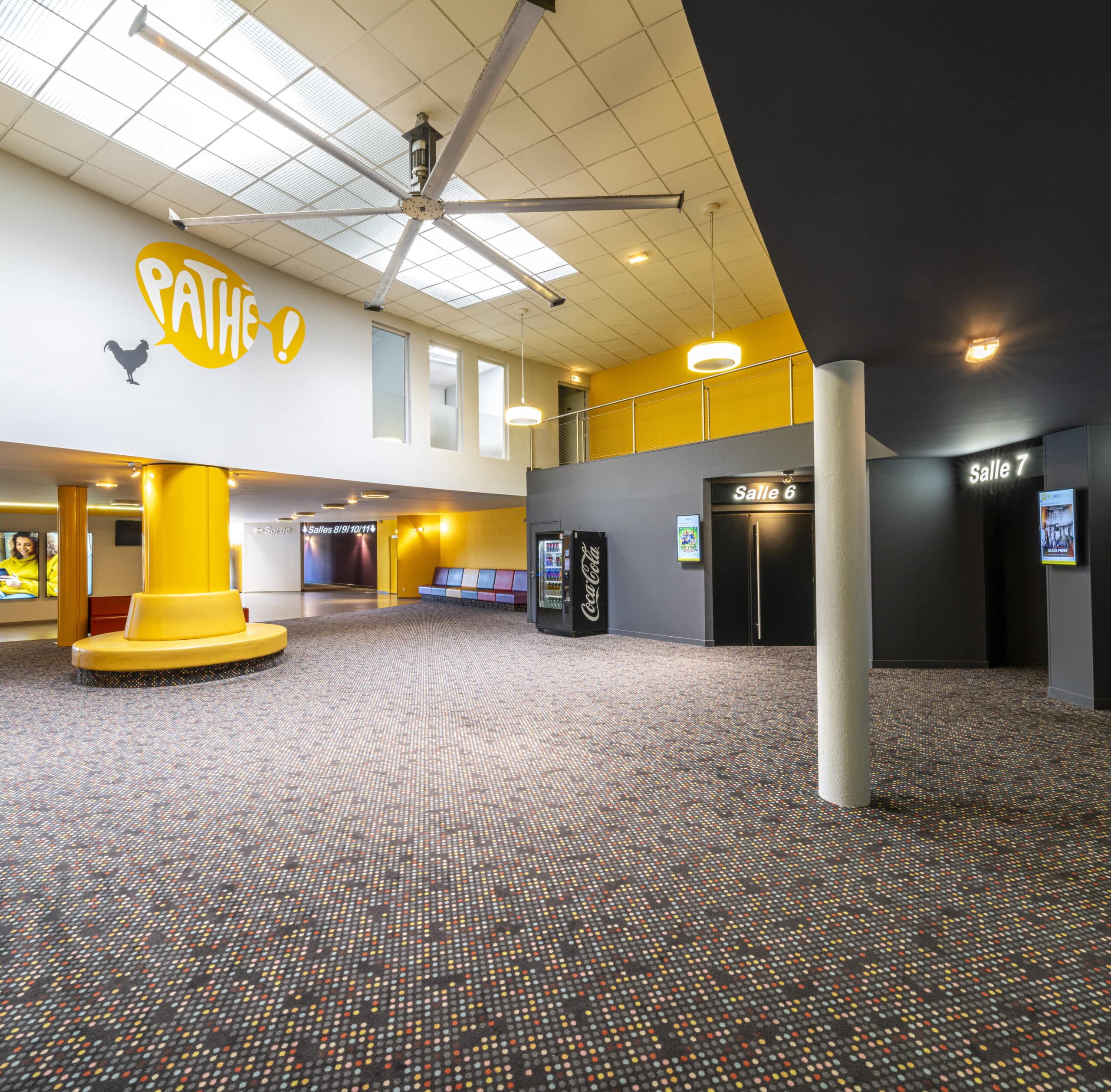 Espace de réception d'un cinéma avec des sièges colorés, un sol à motifs et des colonnes jaunes, entouré par des entrées de salles numérotées.